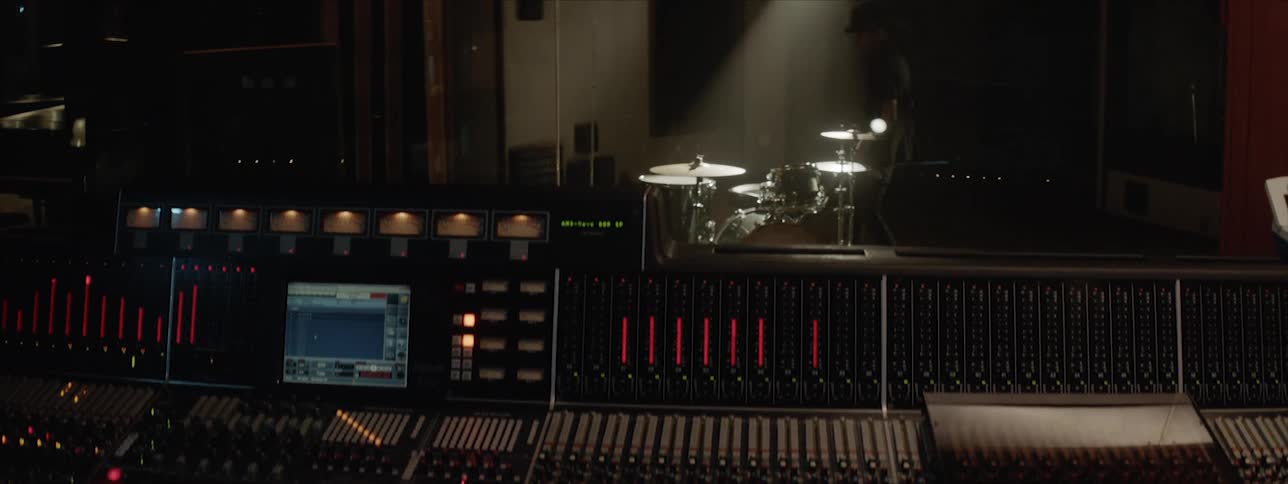 A person with dreadlocks sits at a mixing desk as a drummer wearing a hat sits and plays drums in a studio booth