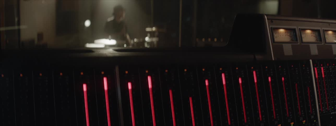 A mixing console displays settings and signals while a drummer wearing a hat sits and plays drums in a studio booth