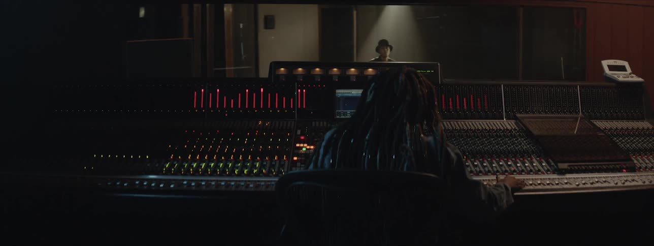A man plays drums in a recording studio while a person works the machines outside
