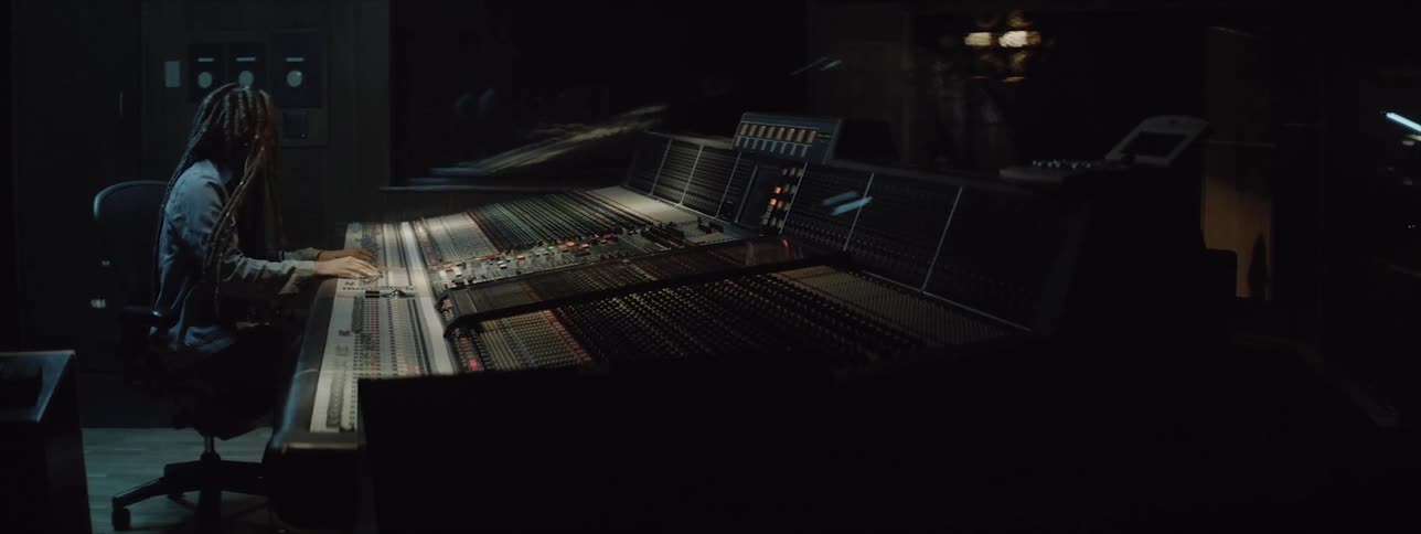 A woman with long dreadlocks sits at an enormous audio mixing board in music studio while nodding her head to the music