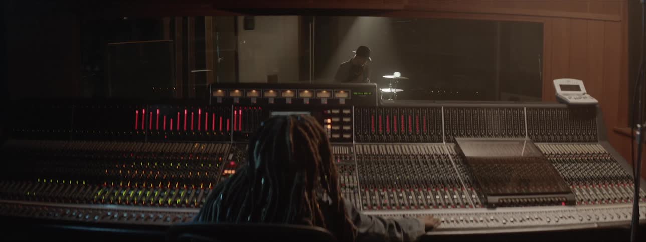A person with dreadlocks sits at a mixing desk as a drummer wearing a hat sits and plays drums in a studio booth