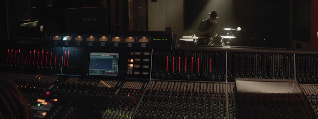 A man sits playing a drum set in a darkened music studio while a female sits at an enormous audio mixing board