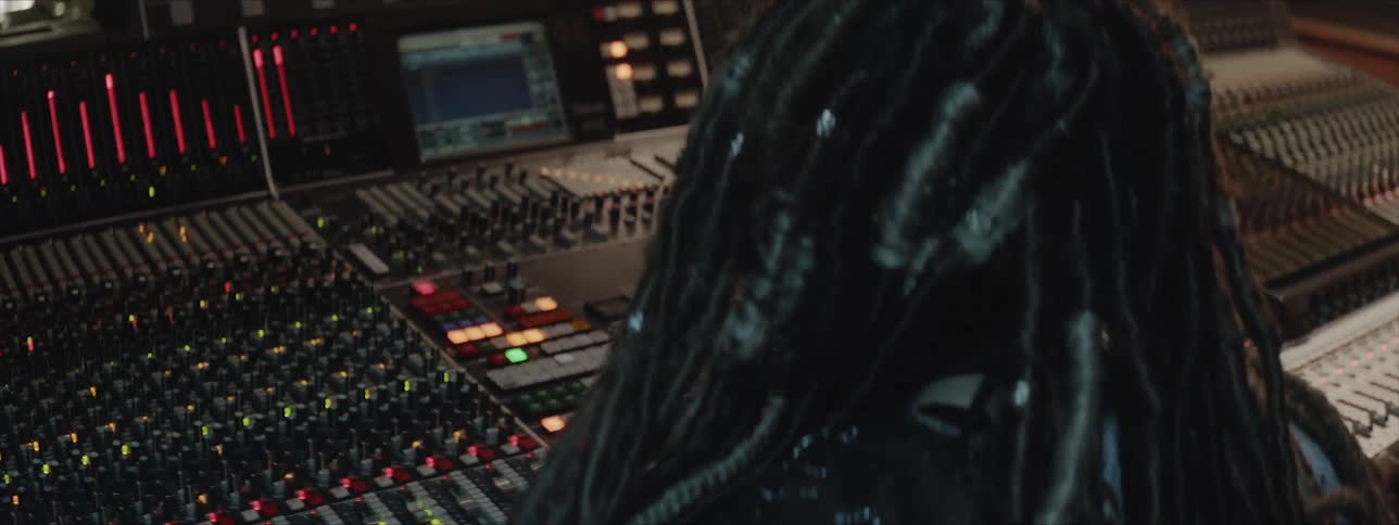 A person with dreadlocks sits at a mixing desk and edits settings