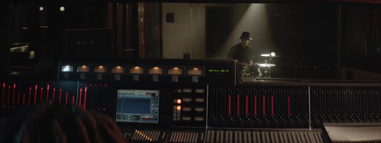 A drummer drumming in a recording studio as a man operates the studio equipment