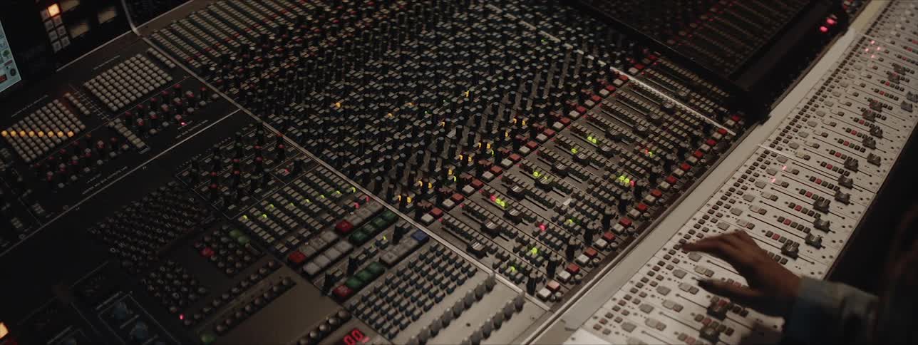 A black woman with long dreadlocks leans over an incredibly large audio mixing board as she makes adjustments to some of the dials