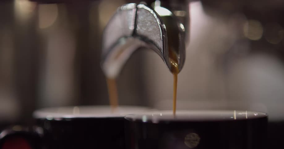 A metallic double spout is seen up close as espresso flows from it into two separate glass espresso cups