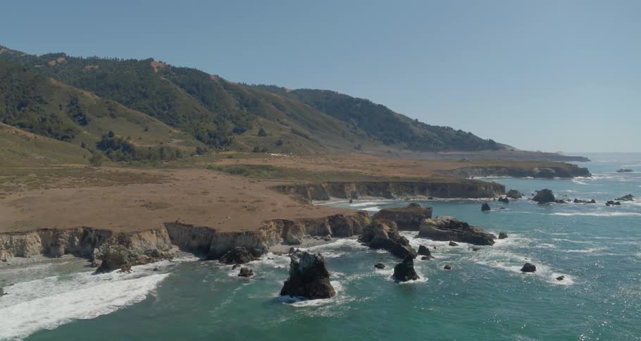 A coastline contains grassy fields that give way to steep cliffs that drop off into the ocean below on a sunny day