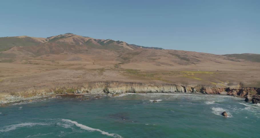 Ocean waves wash up and crash upon the cliffs of this coastal shoreline as grass covered hills can be seen in the background