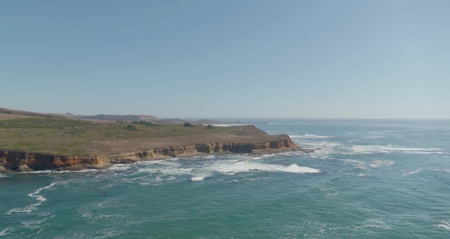 Large sets of waves roll across the ocean surface and waves crash against the cliffs lining a coastline