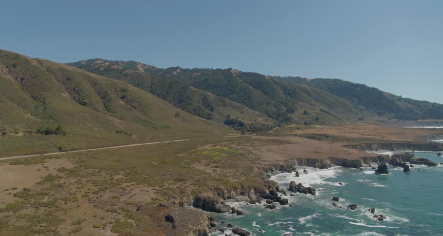 A number of cars speed along a narrow roadway that is near a rocky ocean shoreline and there are large mountains in the background