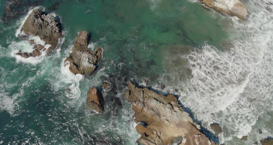 Numerous waves wash over the shallows of a rocky coastal area and crash against the rocks producing very foamy waters