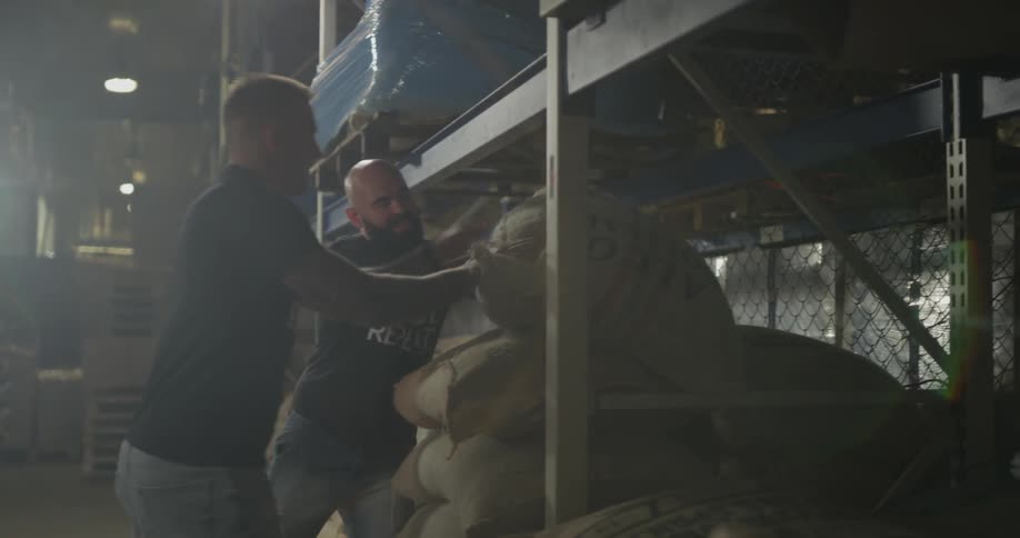 Two men lift a large heavy bag onto a pile of other large bags