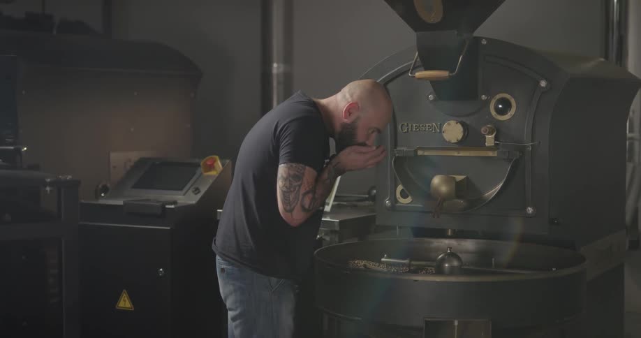 A bald white man standing next to an industrial coffee grinder smelling the coffee beans