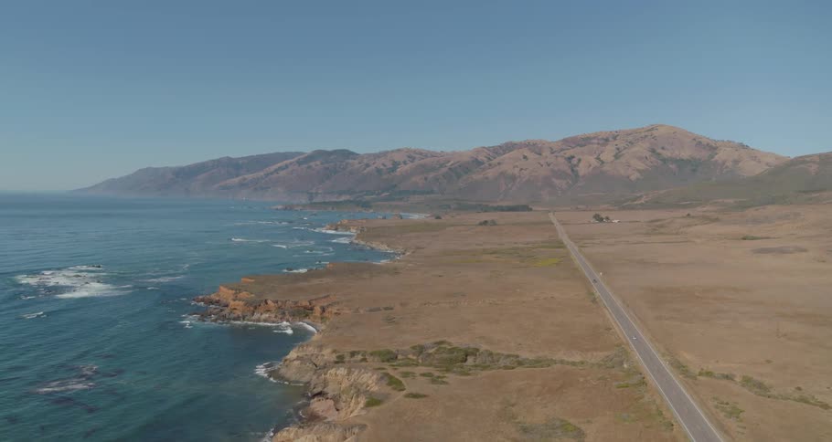 The ocean breaking on a rocky coast with a highway nearby and hills in the distance