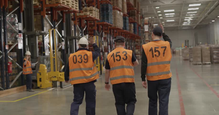 Three men with orange vest walking in a warehouse waving hello to a woman