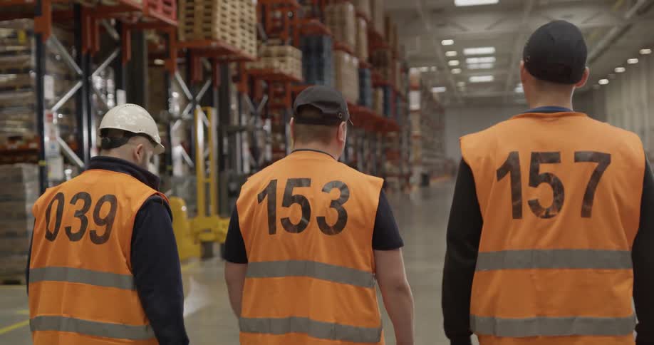 Three men wearing safety vests walk in a factory and wave