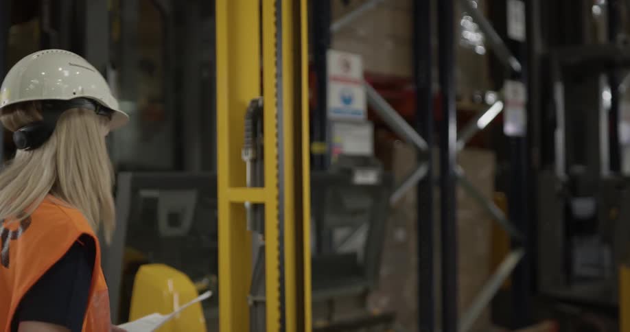 A woman wearing a hard hat is reading a piece of paper then turns to step in a piece of equipment