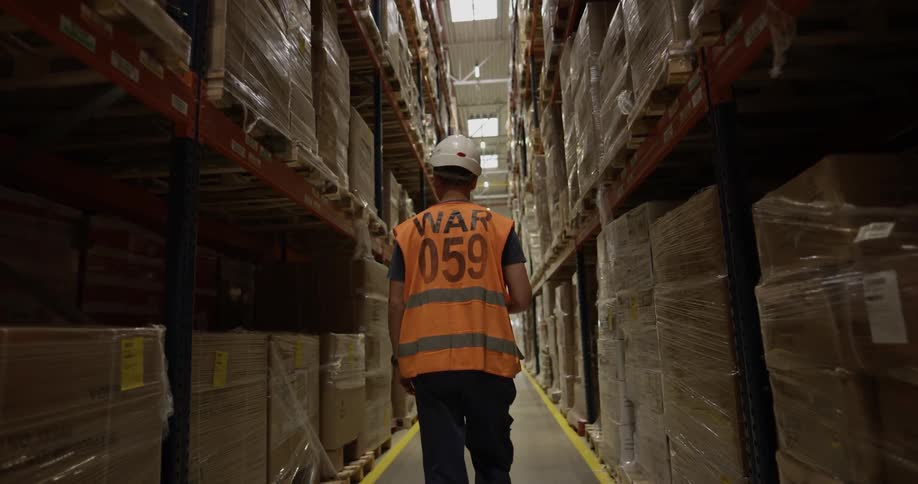 Man wearing a safety vest and helmet walks in a path between filled shelves