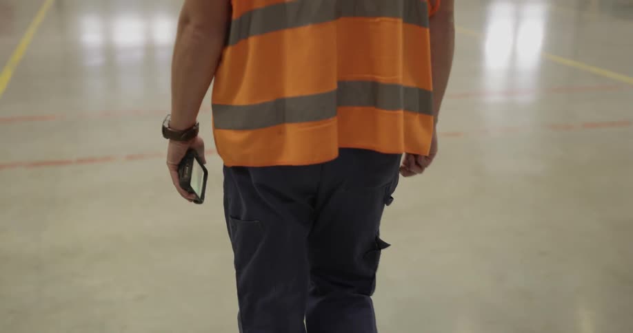 A man wearing a safety vest over a tee shirt and carrying a cell phone walks on a polished warehouse floor
