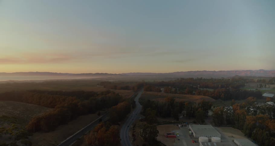 An aerial view shoes plains and forests transected by a long road that are bordered by a number of industrial buildings and facilities