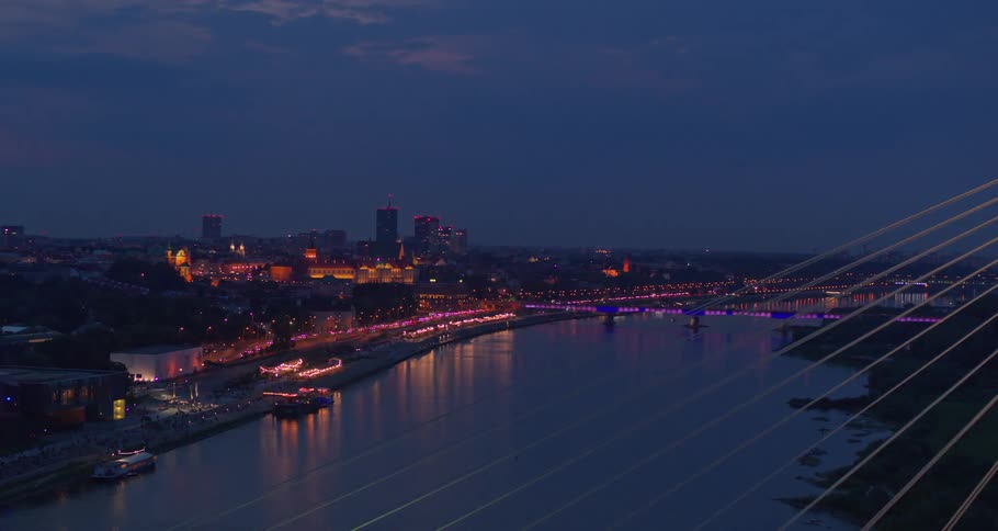 Cables from on bridge stand over the water as purple and blue lights glow in a city skyline and traffic at night