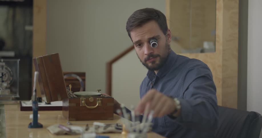 A man putting on a loupe and picking up a screwdriver to work on a device in a wooden box