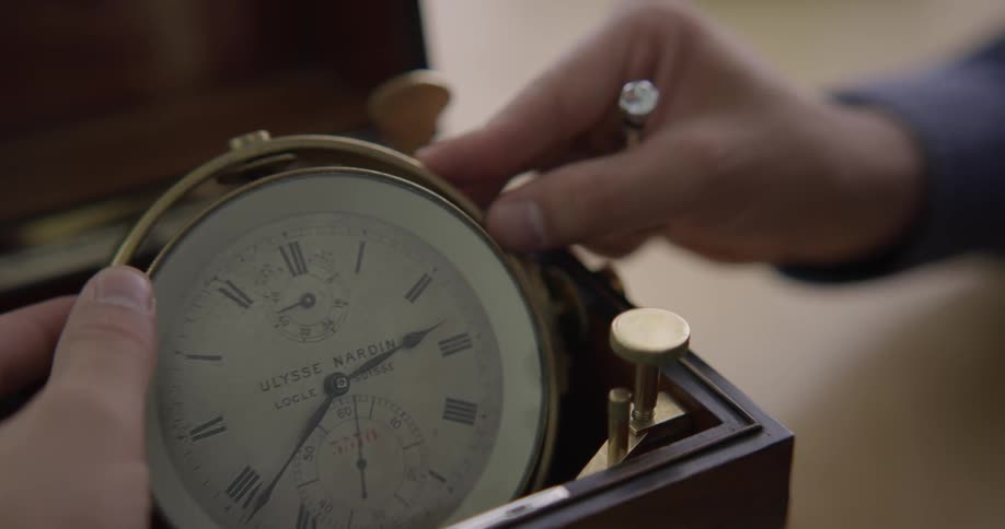 A man turns and examines a ships clock in a wooden case while holding a repair tool in his hand