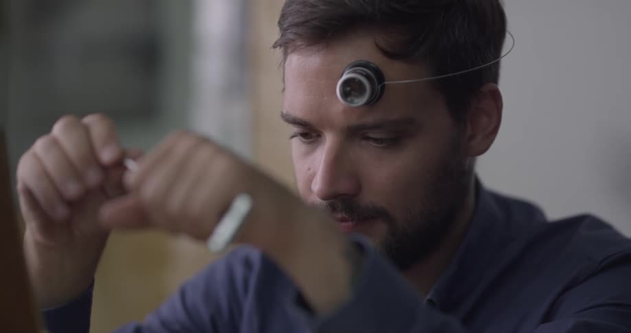 A man with a loupe resting on his forehead fiddling with a tool while lost in thought