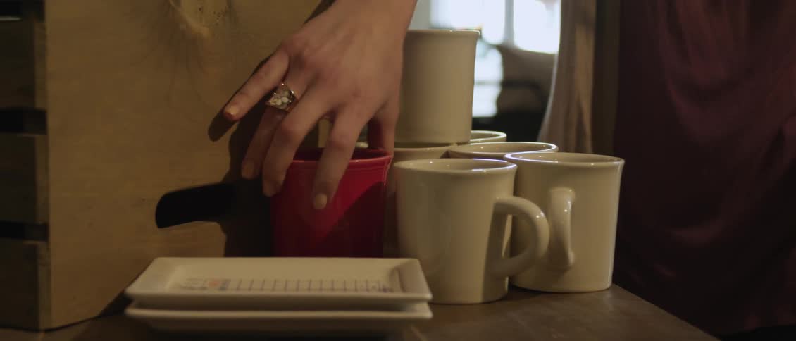 A woman approaches a group of coffee cups on a counter and picks the one red cup and walks away