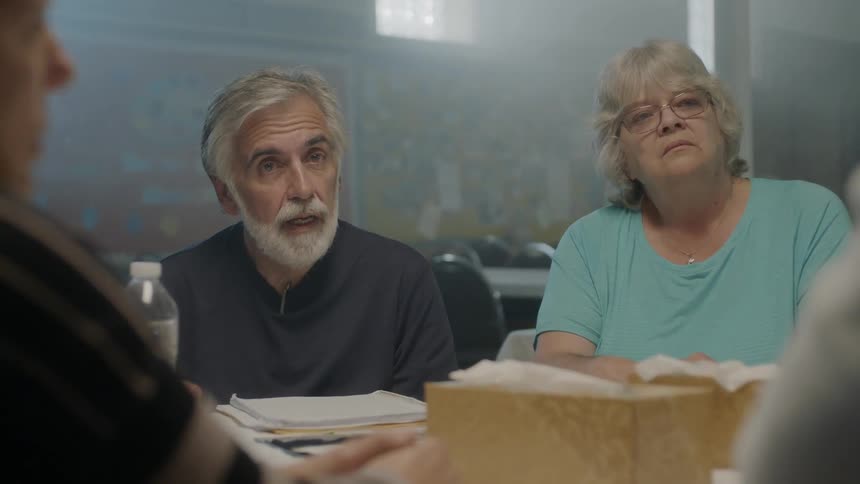 An older man and woman sitting at a table stare at others around them