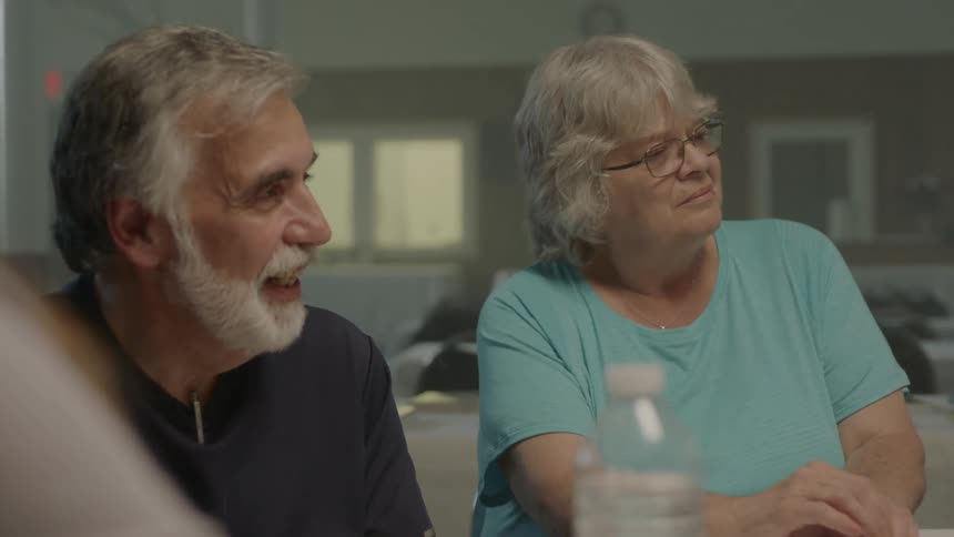 A group of older people sitting around a table listening to a woman in glasses speak