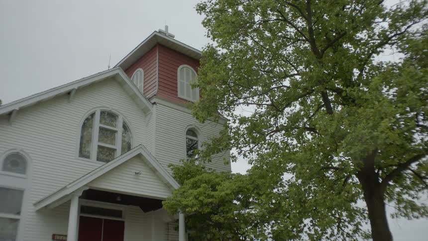 A nice wooden church stands adjacent to a tall tree which has its leafs rustling gently in the wind on a cloudy day