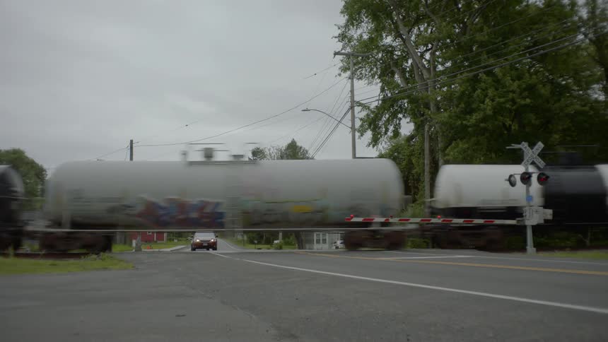 At a train crossing in a small town a few cars wait as a long freight train with many freight cars passes by