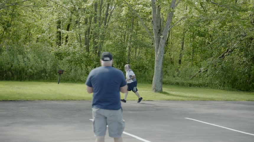 An older man is jogging along the edge of a forested area over a paved parking lot as he participates in a recreational race