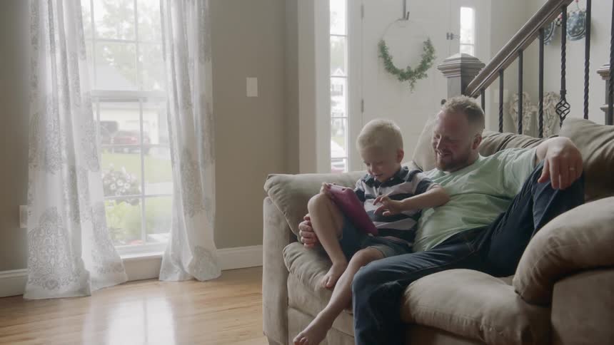 A father and son sit on the couch looking at a tablet