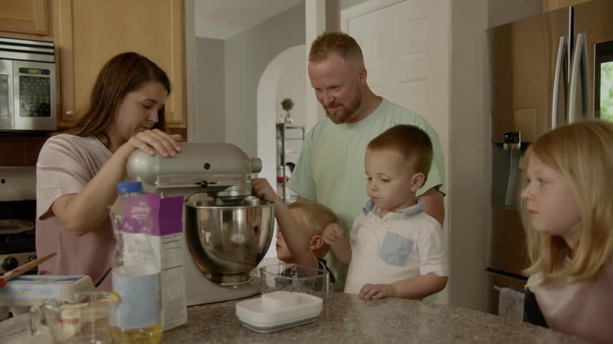 Parents and three blonde children gathered around the kitchen counter as mom uses a stand mixer