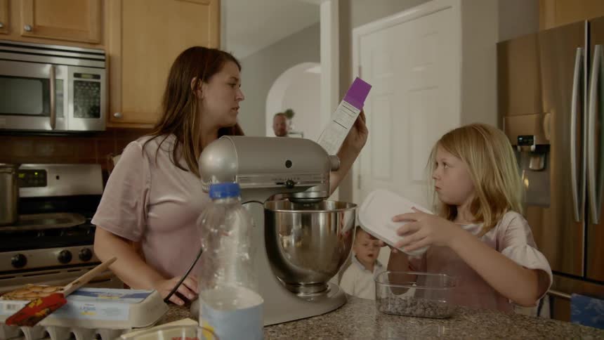 A dad and two young sons walk into the kitchen where a mom and daughter are mixing brownie batter in a mixer