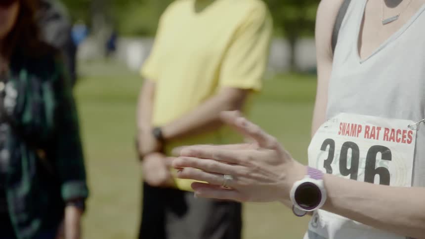 A woman wearing a runners number claps and cheers on the other runners on a sunny and windy day