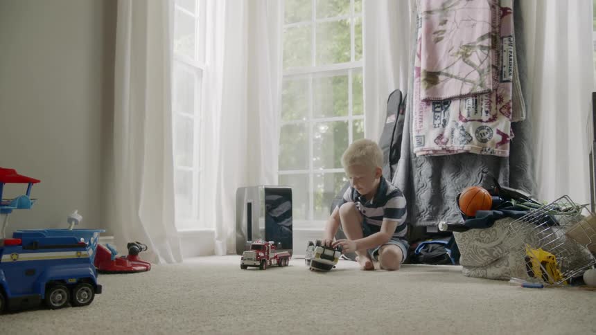 A young boy sits indoors and plays with a toy truck and then another young boy runs in and begins to play besides him