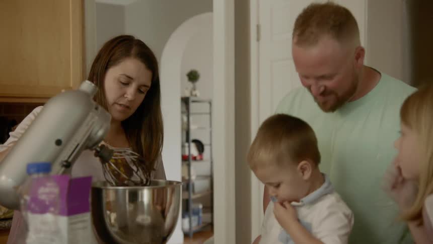 A family standing around a mixer in the kitchen as the mother pokes the content with a wooden spoon