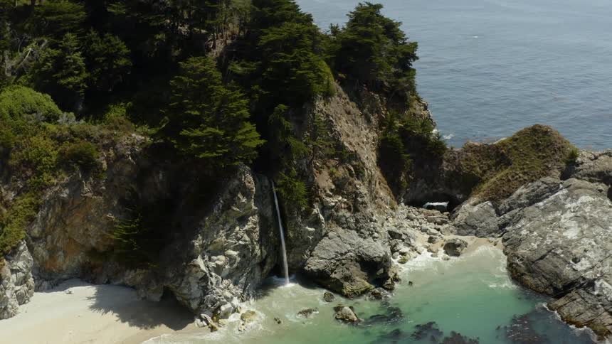 A waterfall sprays water on a beach in a cove