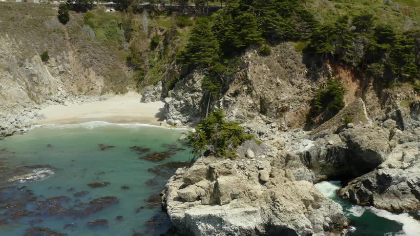 Trees grow on rocks overlooking a sandy beach cove with waves splashing on shore