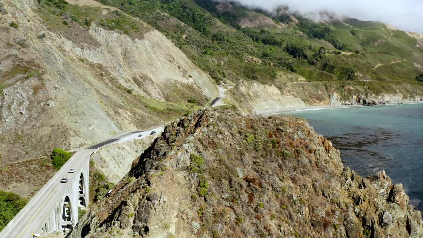Birds eye view of cars traveling across a bridge the water and fog above the hills