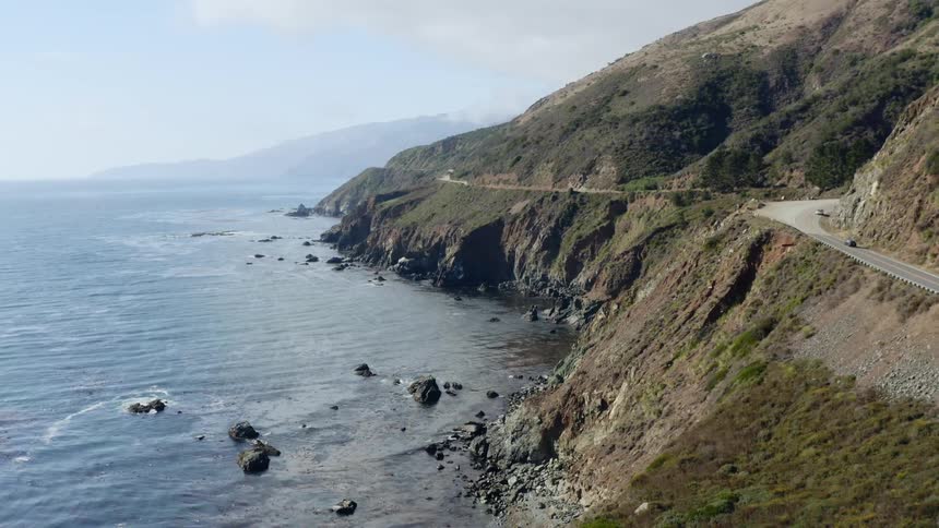 Cars travel on a winding road above the ocean on a blue sky day