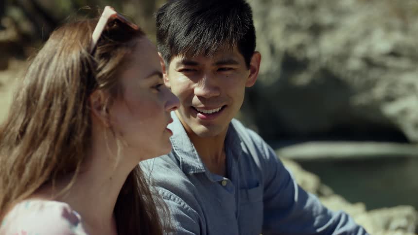 A man smiles and nods as his girlfriend talks and gestures while the wind blows her long hair