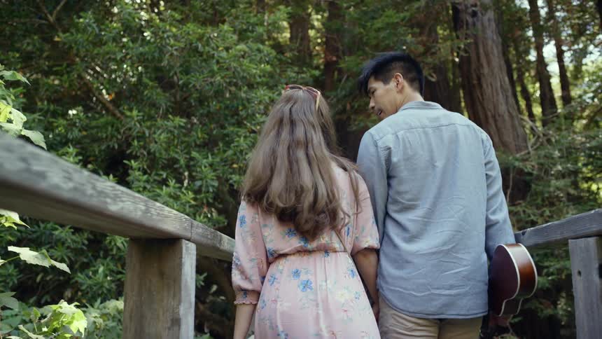 A man carries a guitar and walks with a woman through a forest trail