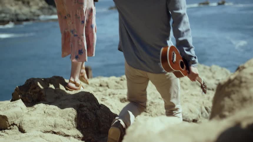 A young man carrying a ukulele holding hands with a young woman carrying a notepad as they cross a boulder on the ocean coast