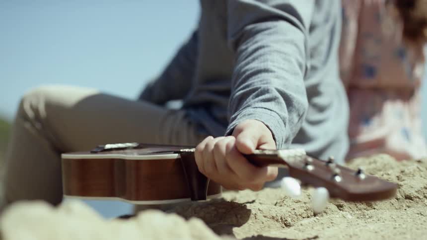 A man reaches back and picks up a ukulele from the rock beside him
