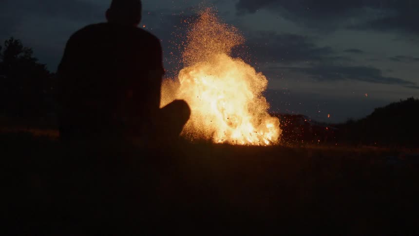 The silhouette of a person sitting cross legged on the grass watching a campfire explode and send flames and sparks into the night sky