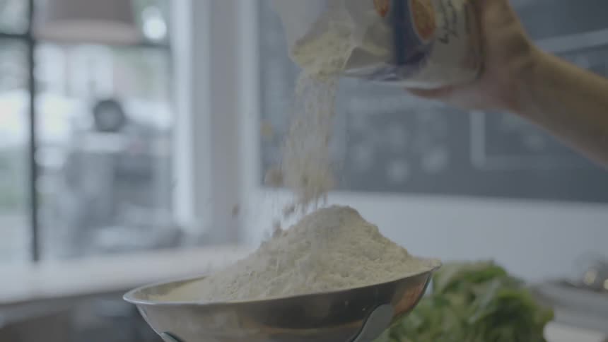 A hand pouring flour from a white bag into a silver bowl